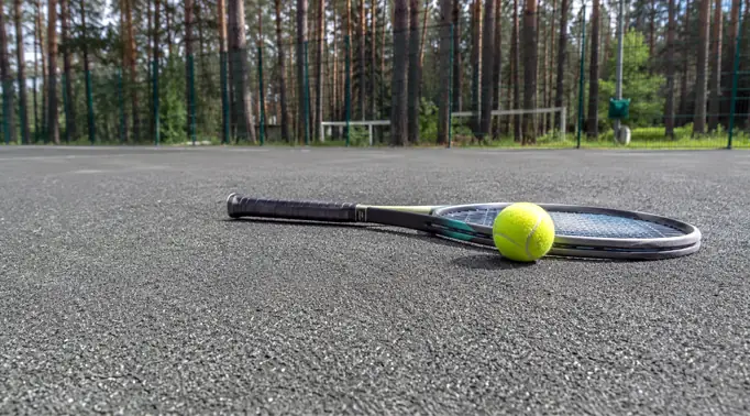 Фотография грунтового теннисного корта: ракетка и желтый мяч лежат на ровном сером покрытии из мелкого гранитного отсева, которое отлично пропускает воду